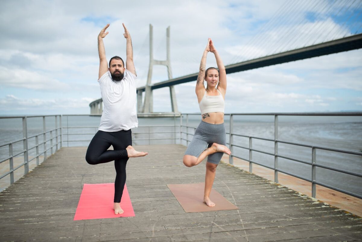 our-stories Two adults practicing yoga on mats outdoors with a bridge backdrop, showcasing healthy lifestyle.
